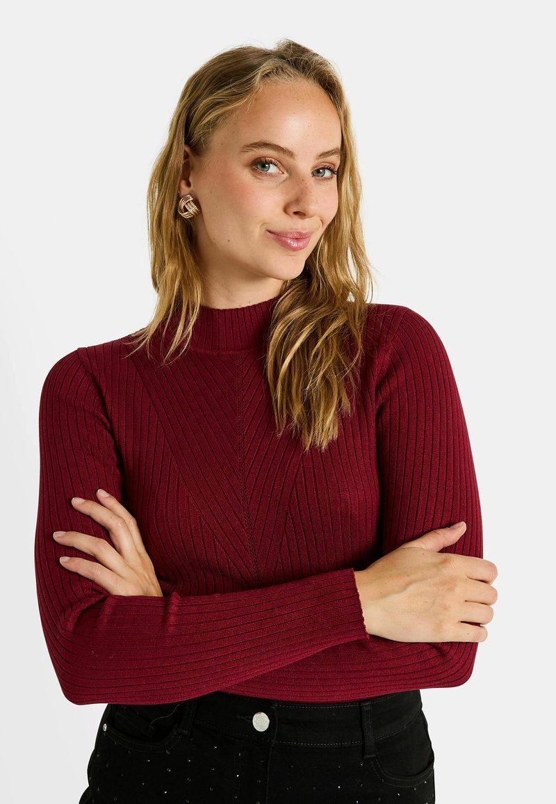 Pull en maille côtelée bordeaux avec un col montant, présentant des rayures en tricot verticales et des manches longues, associé à un pantalon sombre.