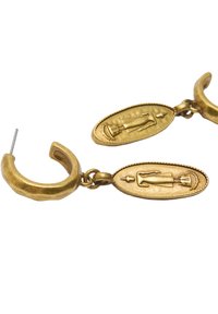 Mosiężne kolczyki z designem na haczyk, z owalnymi charmsami z wygrawerowaną postacią, prezentujące teksturalne wykończenie i metaliczny blask.