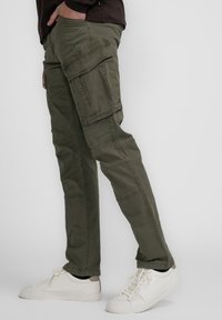 Pantalons cargo en coton vert olive, présentant plusieurs poches, des coutures détaillées et une coupe fuselée. Associés à des baskets blanches.
