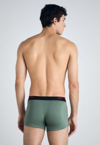 Grüne Boxer Shorts mit schwarzfarbenem Bund und rotem "HUGO"-Logo. Das Design ist eng anliegend, hat eine glatte Textur und einen mittelhohen Schnitt.