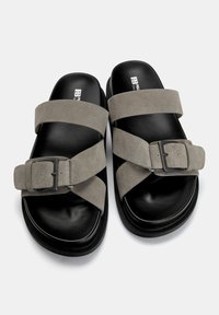 I sandali slide in suede grigio presentano due ampie cinghie con una fibbia rettangolare, poggiando su una suola nera testurizzata. Logo stampato sulla soletta.