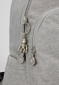 Šedý látkový batoh se dvěma zipovými kapsami, stříbrnými hardware, přívěskem a texturovanými detaily.