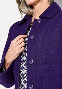 Cappotto di lana viola con colletto, dotato di quattro grandi bottoni e una tasca frontale. Indossato sopra un tessuto a motivi con forme geometriche in bianco e nero.