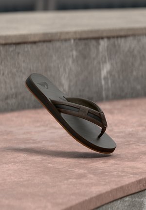 Braune und schwarze Flip-Flop-Sandale, die über einer rosa Steinoberfläche mit grauer Steinwand im Hintergrund schwebt.