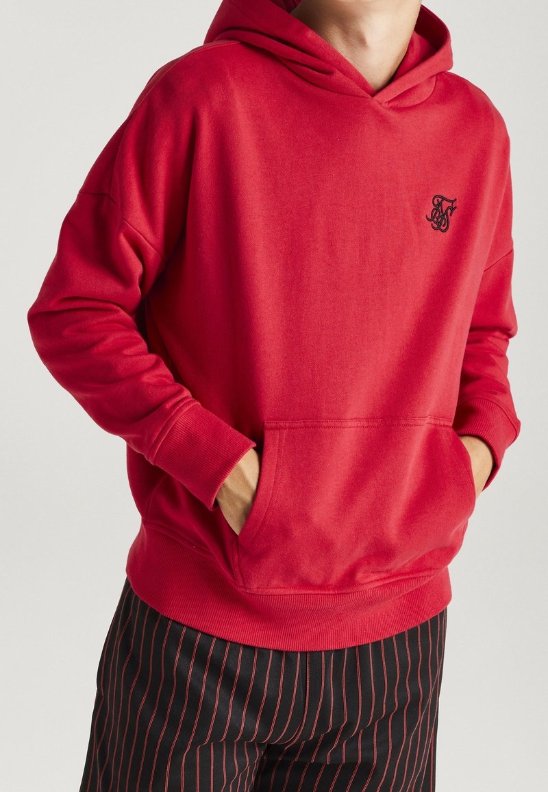 Hoodie rouge avec poche avant et petit logo noir sur la poitrine, porté par une personne les mains dans la poche, assorti à un pantalon noir avec des rayures rouges.