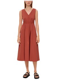 Roestkleurige mouwloze jurk met een diepe V-hals, geplooide taille en volle rok. Gemaakt in combinatie met zwarte sandaaltjes met banden.