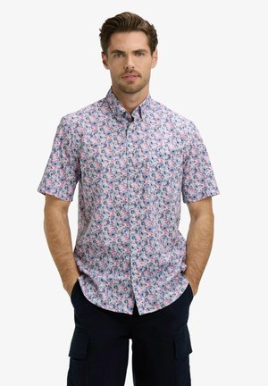Homme portant une chemise à manches courtes avec un motif floral rose et bleu et un pantalon cargo foncé, debout les mains dans les poches.