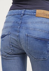 Vaqueros de mezclilla azul con una cintura de altura media, que presentan dos bolsillos traseros y costuras visibles. La tela parece suave con una ligera textura desgastada.