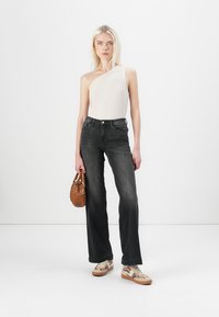 One-shoulder, fitted, light beige top made of metallic fabric; high-waisted dark gray flared jeans; brown handbag; patterned sneakers.