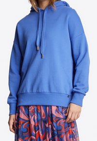 Sudadera azul con capucha de corte holgado, con cordones y puños acanalados. Combinada con una falda estampada y colorida para crear contraste.