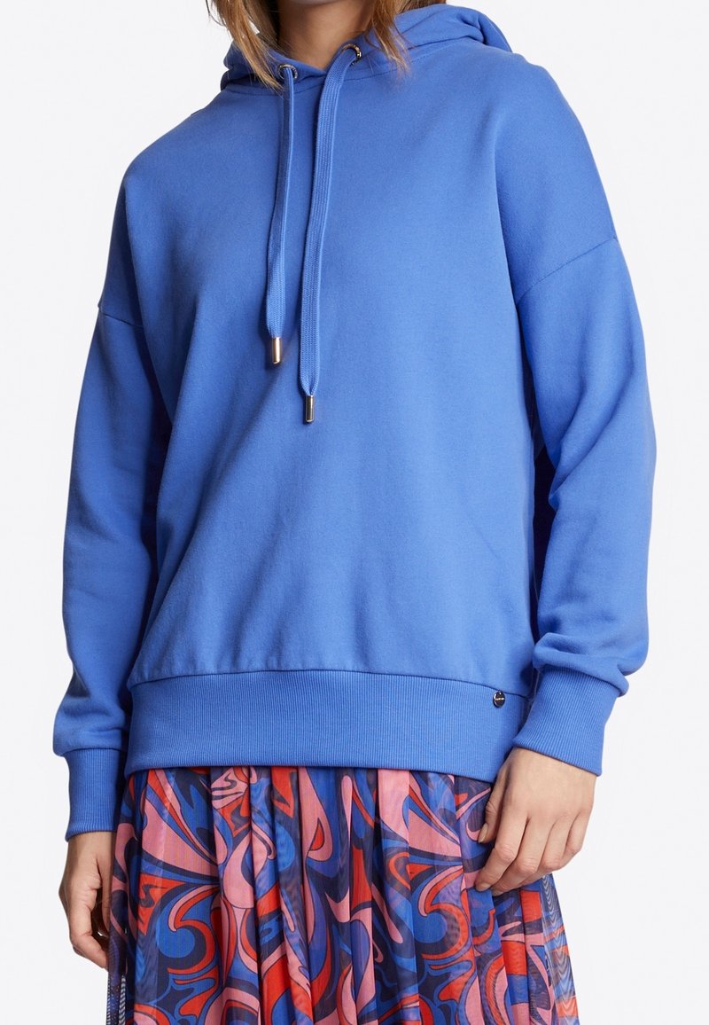 Sudadera azul con capucha de corte holgado, con cordones y puños acanalados. Combinada con una falda estampada y colorida para crear contraste.