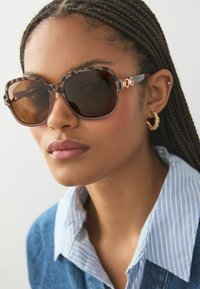 Braune Schildpatt-Sonnenbrille mit dunklen Gläsern, rechteckiger Form und dekorativen goldenen Verzierungen. Getragen mit einem blau gestreiften Hemd mit Kragen.