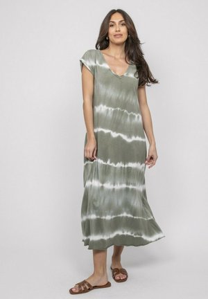 Femme aux cheveux longs et foncés portant une robe midi sans manches tie-dye verte et blanche et des sandales marron, debout devant un fond clair uni.