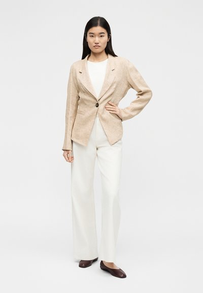 Γυναίκα που στέκεται με το χέρι στη μέση, φορώντας ένα μπεζ σακάκι με κουμπιά, λευκό παντελόνι, λευκό τοπ και σκούρα καφέ παπούτσια σε απλό φόντο.