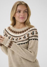 Pull en tricot beige avec un empiècement à motif en crème, marron et noir. Il présente des manches longues et des poignets côtelés. Texture douce, col rond.
