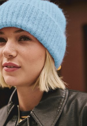 Jeune femme aux cheveux blonds courts porte un bonnet tricoté bleu clair, une boucle d'oreille en or et une veste en cuir noire, regardant légèrement sur le côté.