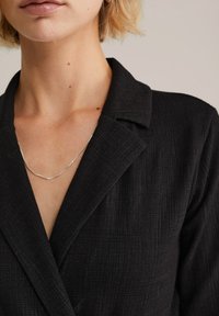 Melna jaka ar faktūru, kurai ir piegriezta apkakle un V veida kakla izgriezums. Sudraba ķēdes kaklasprādze ir redzama pie izgriezuma.