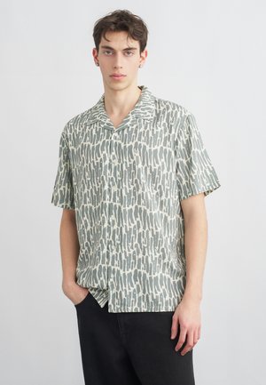 Mladý muž nosiaci košeľu s krátkym rukávom so zapínaním na gombíky, s abstraktným sivým a bielym zvislým vzorom, čierne nohavice, stojaci s jednou rukou v vrecku.