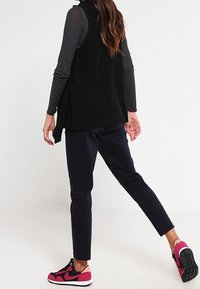 Svart stickad väst över en mörk långärmad skjorta, kombinerad med marinblå byxor och rosa- och svarta sneakers. Strukturerad tyg, avslappnad stil.