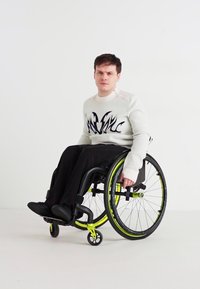 Ένα άτομο σε αναπηρικό καροτσάκι φοράει ένα λευκό φούτερ με μαύρα γραφικά σχέδια και μαύρο παντελόνι. Το αναπηρικό καροτσάκι έχει νιον πράσινες λεπτομέρειες.