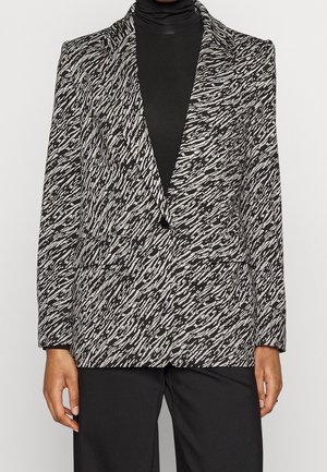 Schwarz-weißer Blazer mit Muster und Einknopfverschluss, getragen über einem schwarzen Rollkragenpullover und kombiniert mit schwarzen Hosen.