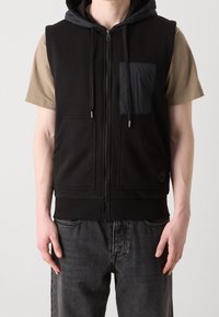 Muž v černé neformální bezrukávové mikině se zipem a kapsami vpředu, pod kterou má béžové tričko s krátkým rukávem a tmavě šedé džíny.