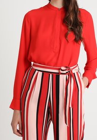 Röd långärmad blus med knäppning i halsen. Kombineras med högmidjade randiga byxor i rosa, röd och svart, med knytband i midjan.