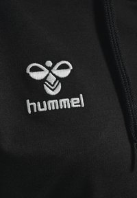 Juodas audinys su siuvinėtu baltu logotipu, kuris vaizduoja stilizuotą bičių ir tekstą „hummel“ po juo. Audinio tekstūra atrodo lygi ir matinė.