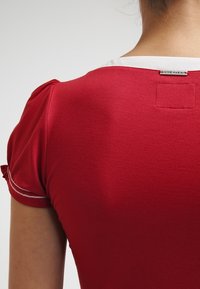 Mujer que lleva una camiseta de manga corta roja con ribetes blancos y una etiqueta visible de "Vive Maria" en el cuello de la parte trasera.
