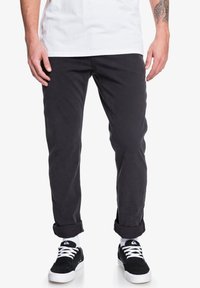 Pantalon en coton noir, coupe slim, avec des ourlets retroussés, associé à des baskets noires. La texture semble lisse, sans motifs ni ornements visibles.