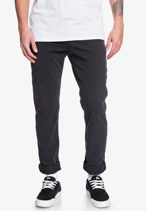 Czarne bawełniane spodnie, slim fit, z podwiniętymi nogawkami, w połączeniu z czarnymi sneakersami. Tekstura wydaje się gładka, bez widocznych wzorów czy ozdób.