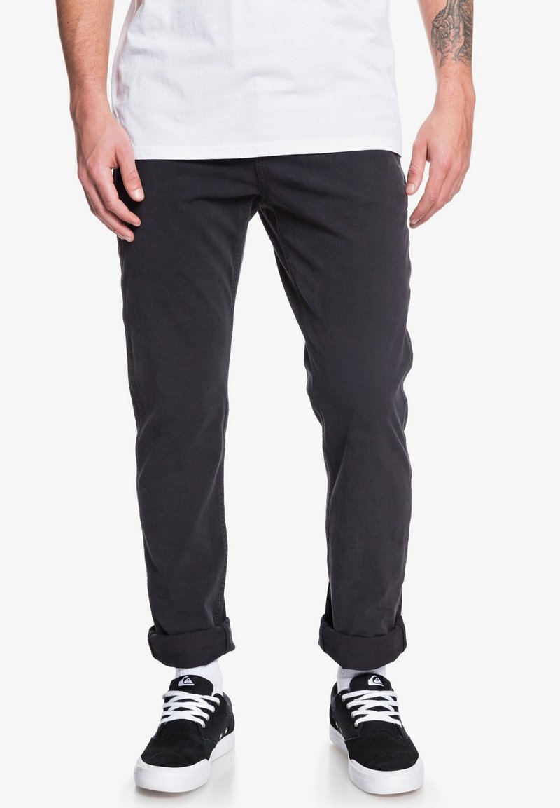 Pantalon en coton noir, coupe slim, avec des ourlets retroussés, associé à des baskets noires. La texture semble lisse, sans motifs ni ornements visibles.