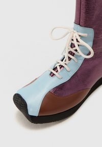 Botín multicolor que presenta una parte superior de cuero azul, acentos metálicos morados, secciones de cuero marrón y cordones blancos con una suela negra texturizada.