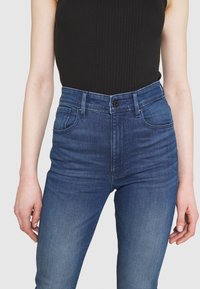 Högmidjade jeans i mörkblå denim med en slät textur, femfickorsdesign och en knappstängning i midjan.