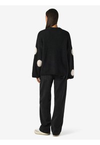 Suéter negro con un acabado texturizado y acentos de pompones blancos en las mangas, combinado con pantalones negros holgados y zapatos informales.