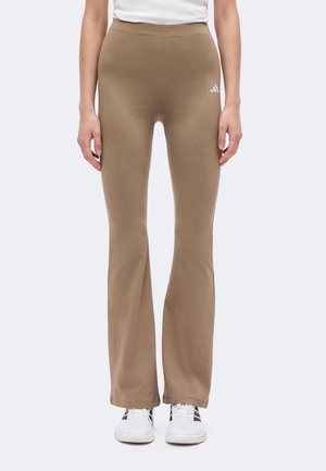 Personne portant un pantalon évasé beige avec le logo Adidas et des baskets blanches, debout devant un fond clair uni.