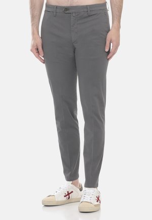 Uomo che indossa pantaloni grigi slim fit e sneakers bianche con logo rosso, in piedi contro uno sfondo grigio chiaro.