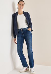 Giacca zip-up blu navy sopra una maglietta bianca a scollo a V, abbinata a jeans blu medio e sneaker bianche con dettagli rossi; in piedi contro uno sfondo chiaro.