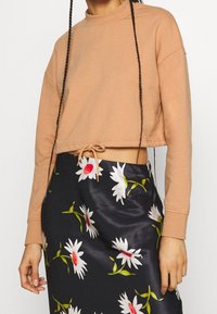 Mjuk, beige kort sweatshirt med hög halsringning och justerbar nederdel, kombinerad med en blommig svart satin kjol med vita och röda blommor.