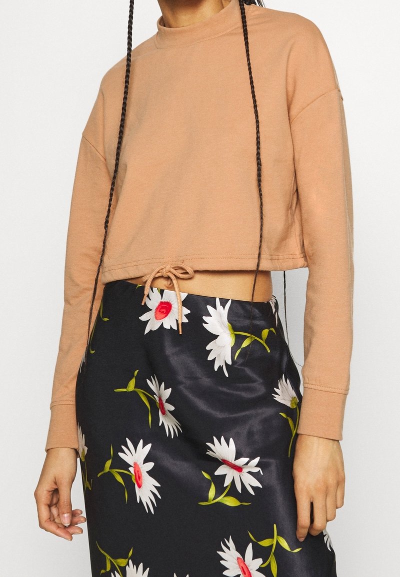 Mjuk, beige kort sweatshirt med hög halsringning och justerbar nederdel, kombinerad med en blommig svart satin kjol med vita och röda blommor.