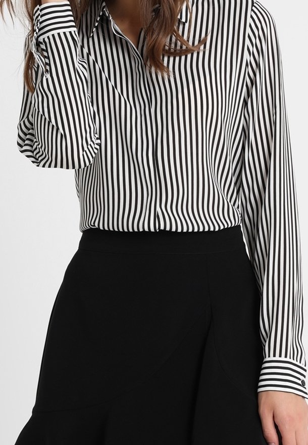 Femme portant une chemise à manches longues rayée verticalement en noir et blanc, rentrée dans une jupe noire taille haute.