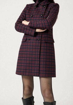 Persona che indossa un cappotto doppiopetto bordeaux e nero a quadri, collant a rete neri e stivaletti neri in pelle, braccia incrociate.