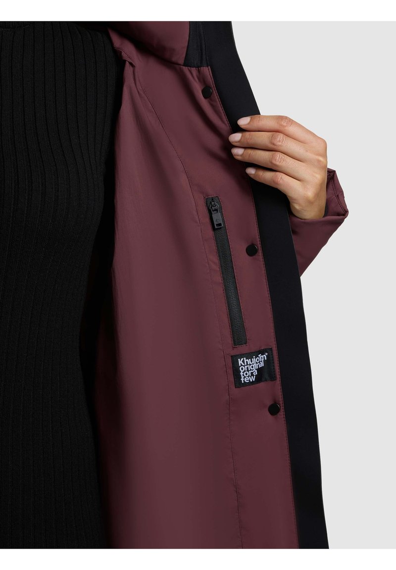 Interiorul jachetei impermeabile burgundy cu accente negre, buzunar cu fermoar și patch cu logo; textură netedă cu închideri tip buton.