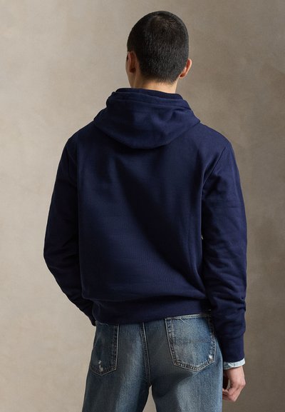 Νavy μπλε φούτερ με κουκούλα και υφασμάτινη υφή, τσέπη καγκουρό και ριμπ μανσέτες, συνδυασμένο με ανοιχτόχρωμο τζιν παντελόνι.