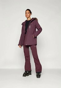 Σακάκι σκι σε σκούρο μοβ ύφασμα με κουκούλα με γούνινη επένδυση, με τσέπες με φερμουάρ, συνδυασμένο με αντίστοιχες παντελόνα σκι και μαύρες μπότες.