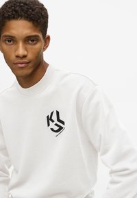 Fiatal férfi fehér pulóverben, fekete KLS logóval és Karl Lagerfeld márkajelzéssel a bal mellkasán, sima fehér háttér előtt.