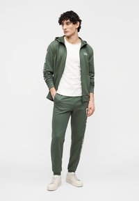 Groene sporthoodie met rits en zakken, gecombineerd met bijpassende joggers. Witte T-shirt eronder en lichtgekleurde sneakers.