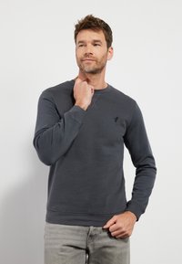 Sudadera gris oscuro con cuello redondo, mangas largas y un pequeño logo en el pecho. Textura suave con puños y dobladillo acanalados.