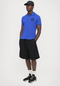 Μπλε πόλο μπλούζα με μαύρο λογότυπο, κοντά μανίκια, μπλε γιακάς, συνδυασμένη με μαύρα πλισέ σορτς και μαύρα αθλητικά παπούτσια με λευκή σόλα.