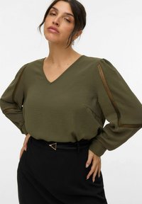 Oliven grøn bluse med V-hals og pufærmer, der har mesh-detaljer. Kombineret med en højtaljet sort nederdel og en triangular bæltespænde.